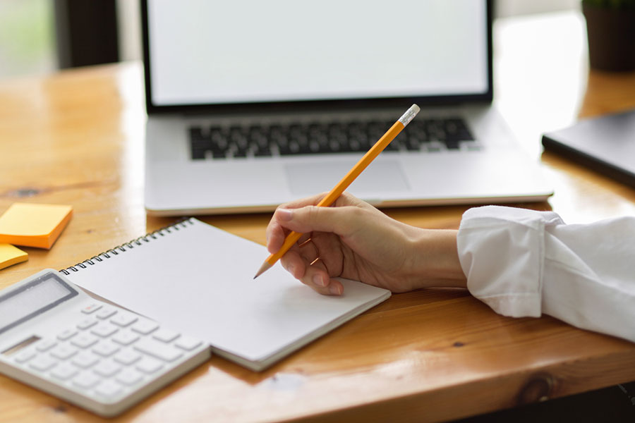 Person writing in notebook with pencil at desk, next to calculator and laptop