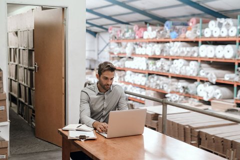 Man working on a laptop in a warehouse with shelves full of rolled stock and packaging materials